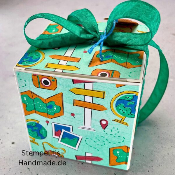 Handdekorierte Fensterschachtel mit Reisethema und grünem Band, Detailansicht des Sichtfensters mit Füllmaterial, Koffer-Motiv auf der Front der Schachtel, Oberseite mit Geschenkanhänger „Reiselust“, Seitliche Ansicht der Box mit Weltkartenmotiv, Geöffnete Box mit Blick auf das Innere, Fensterschachtel als kreative Geschenkverpackung für Reisegutscheine